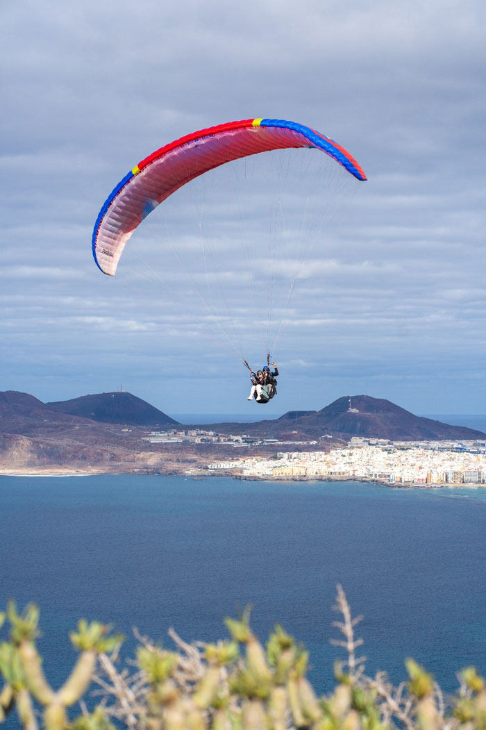 Piloto de parapente en primer plano volando en biplaza con un pasajero, con los acantilados de Gran Canaria como impresionante telón de fondo.